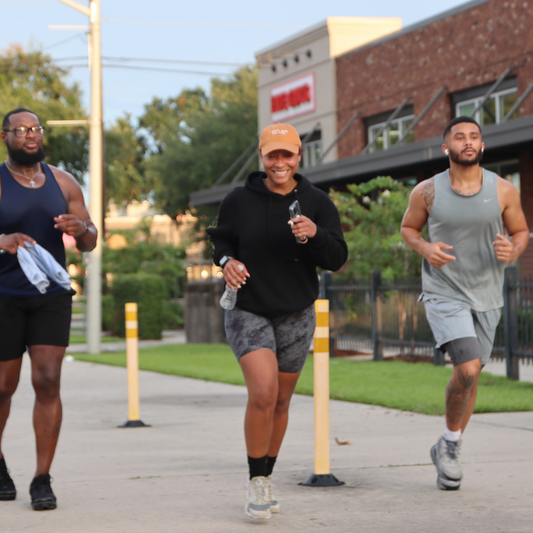 Three runners, two men and one woman, running outdoors during a Nola Miles Club event. The woman smiles while holding a water bottle, and the group runs along a paved path near a building.